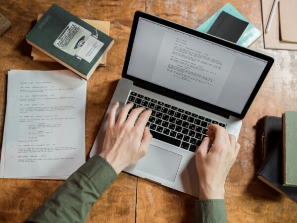 High-angle view of hands typing on a laptop surrounded by books and papers.